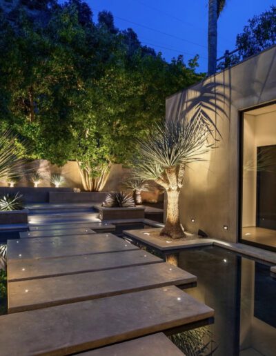 A modern home entrance at dusk with floating stone steps over water, flanked by illuminated palm trees and a landscaped garden.