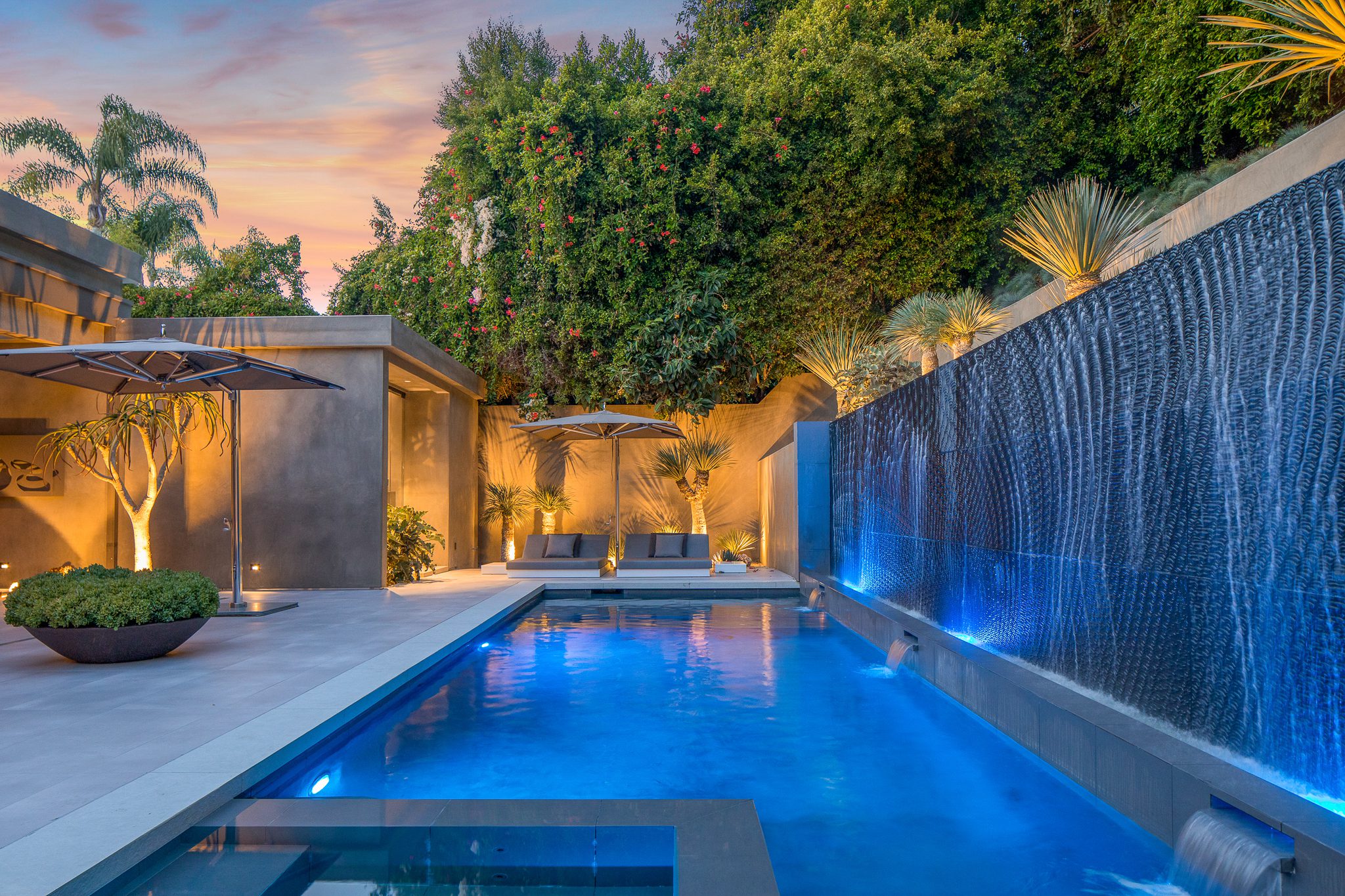 Luxurious backyard pool with a cascading water feature and ambient lighting at dusk.