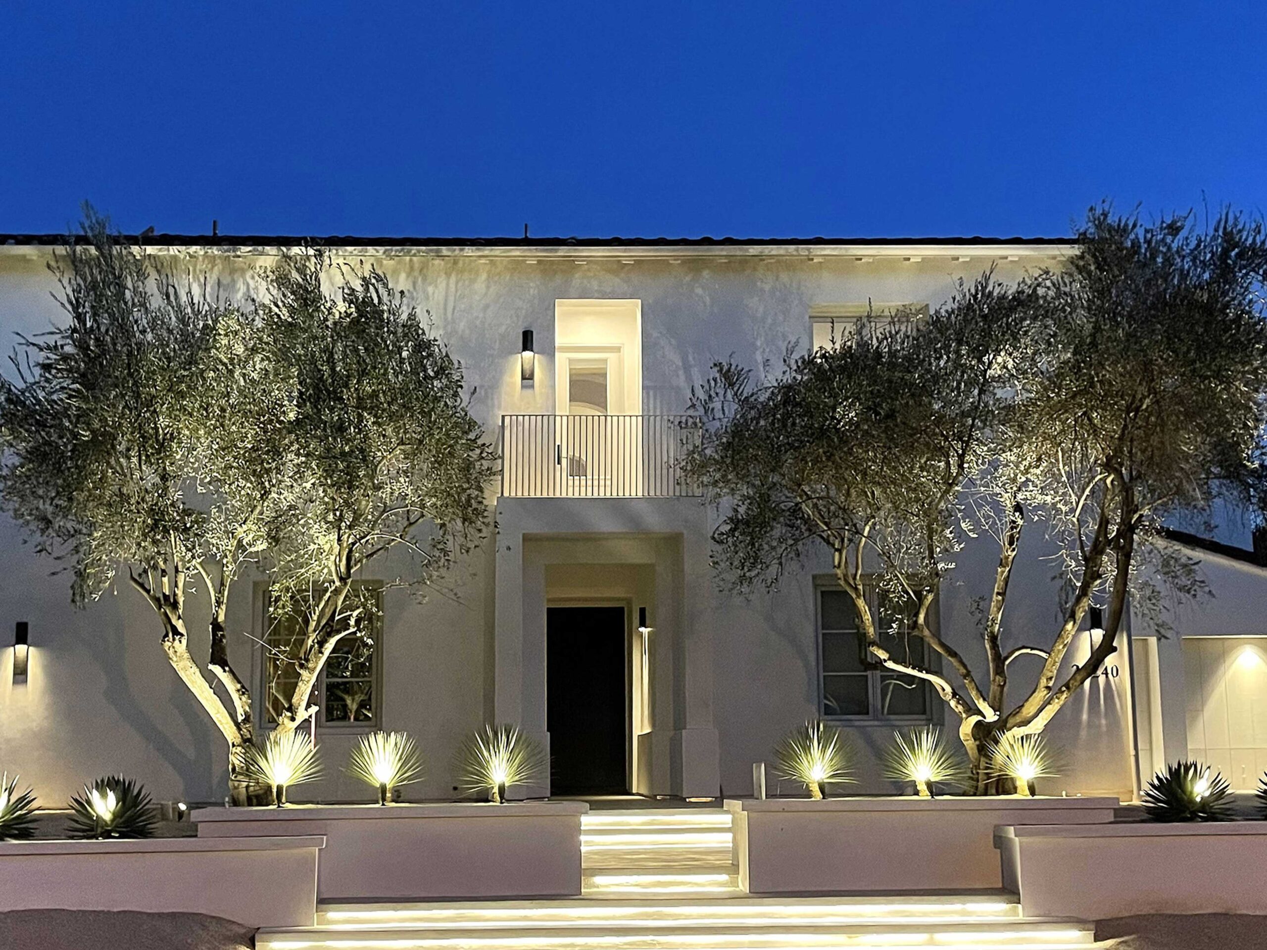 A two-story house at twilight with exterior lighting and olive trees flanking the entrance steps.